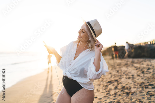 Cheerful female in modern earphones spending summertime at Philippines coastline, happy Caucaisan woman in electronic headphones listening audio book via mobile app during vacations on Tahiti #491891428