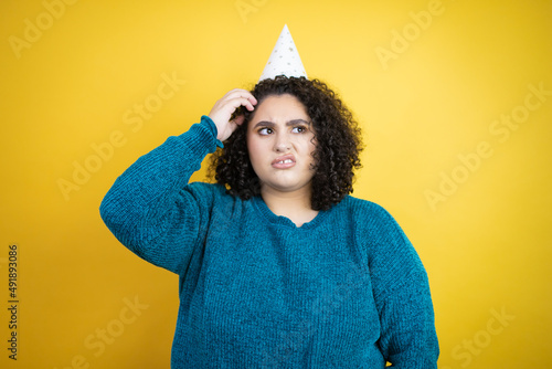 Young beautiful woman wearing a birthday hat over isolated yellow background confuse and wonder about question. Uncertain with doubt, thinking with hand on head © Irene