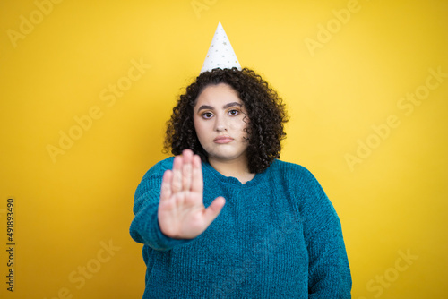 Young beautiful woman wearing a birthday hat over isolated yellow background serious and doing stop sing with palm of the hand. © Irene
