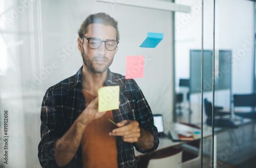 Caucasian employee 30s dressed in smart casual wear pointing on colorful papers with text notes glued on glass board, male entrepreneur in spectacles reading memo stickers working in office interior