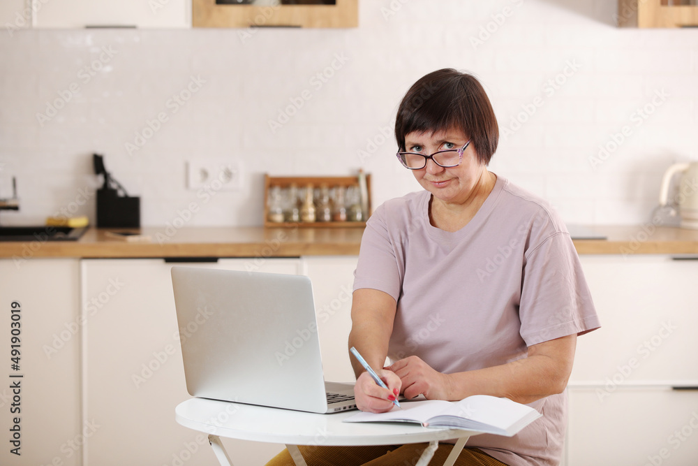 mature old lady working remotely from home office on laptop making notes. Smiling middle aged 50s business woman using computer watching webinar sitting at the table at kitchen and writing mature old lady working remotely from home office on laptop making notes. Smiling middle aged 50s business woman using computer watching webinar sitting at the table at kitchen and writing