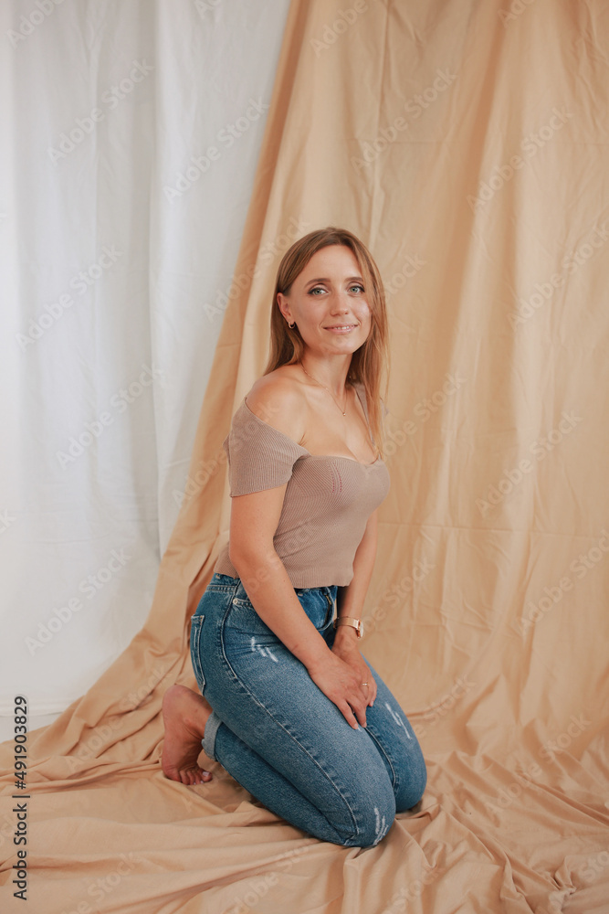 Beautiful Ukrainian girl in a photo studio. Portrait of a cute girl on a beige fabric background. Tanned woman in jeans and beige top. Classic portrait in the studio. Beautiful Ukrainian girl in a photo studio. Portrait of a cute girl on a beige fabric background. Tanned woman in jeans and beige top. Classic portrait in the studio.