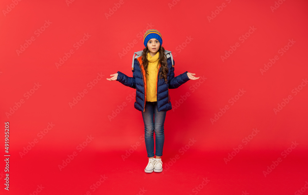 full length of confused child in knitted winter hat and down coat on red background, teenager