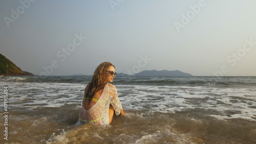 Sexy fit woman in a white tunic on the beach, near the stormy sea. Happy playful blonde with wet curly hair, in sunglasses. Lady tourist enjoying vacation time, walks has fun. Blue swimsuit, pareo. © ivandanru