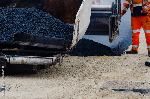 Asphalt paver filled with hot tarmac laying new road surface on new residential housing development site and roadworker operator in orange hi-viz next to it photo