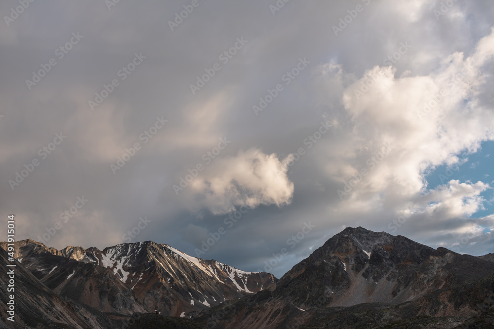 Dramatic mountain landscape with high mountain range with sharp rocky pinnacle under clouds of sunset color in gloomy sky. Dark atmospheric scenery with large mountains with snow at changeable weather