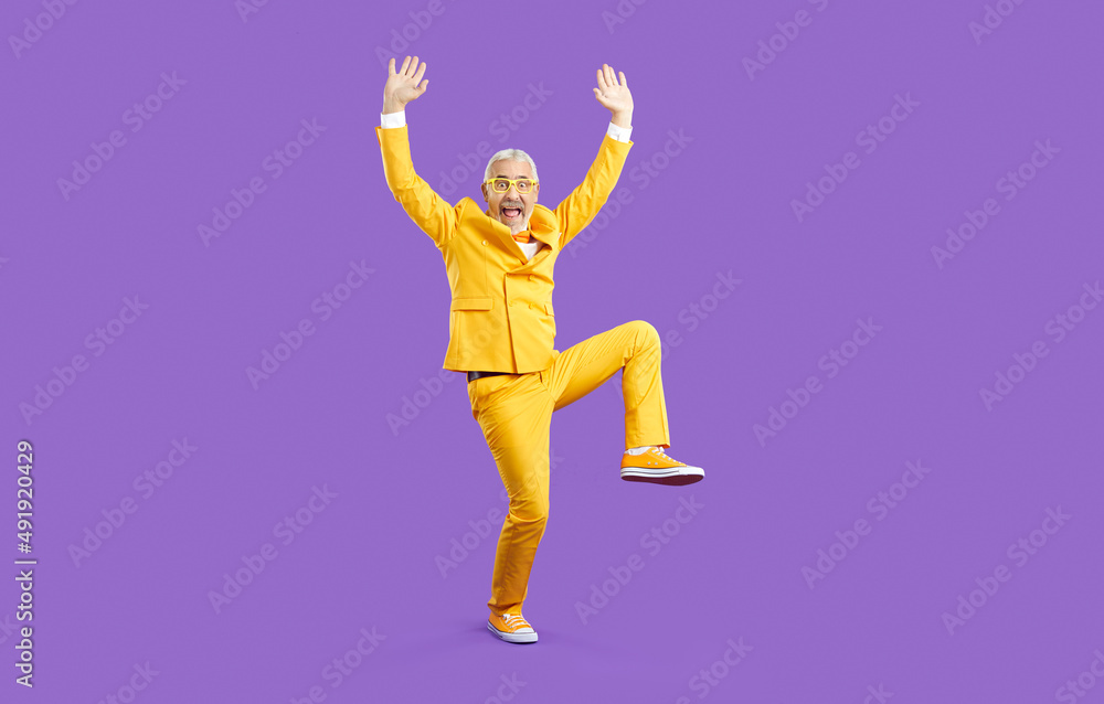 Funny old man having fun and fooling around in the studio. Happy crazy senior man wearing a bright yellow party suit and glasses standing with his hands raised up isolated on a solid purple background