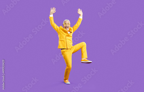 Fototapeta Naklejka Na Ścianę i Meble -  Funny old man having fun and fooling around in the studio. Happy crazy senior man wearing a bright yellow party suit and glasses standing with his hands raised up isolated on a solid purple background