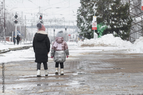 Two sisters of different ages walk along the railway platform with their backs to us, in the winter season. Traveling in winter with children.