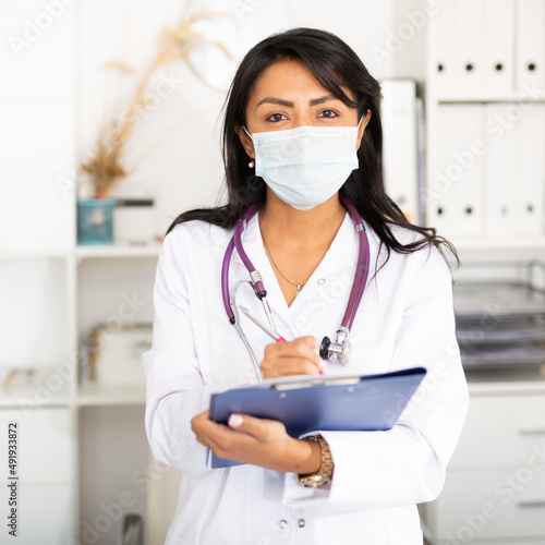 Portrait of latin american female doctor in surgical face mask meeting patient in medical office, filling out medical form at clipboard