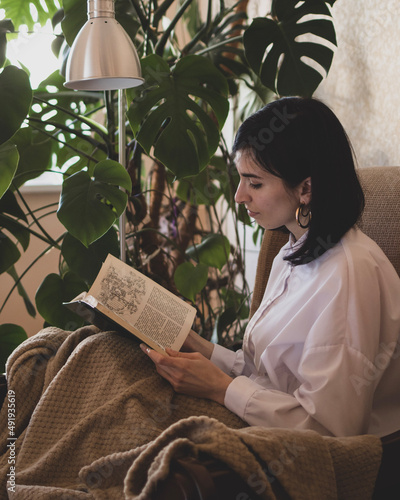 Φωτογραφία Young woman is reading a book and sitting at home in a chair with bedcover