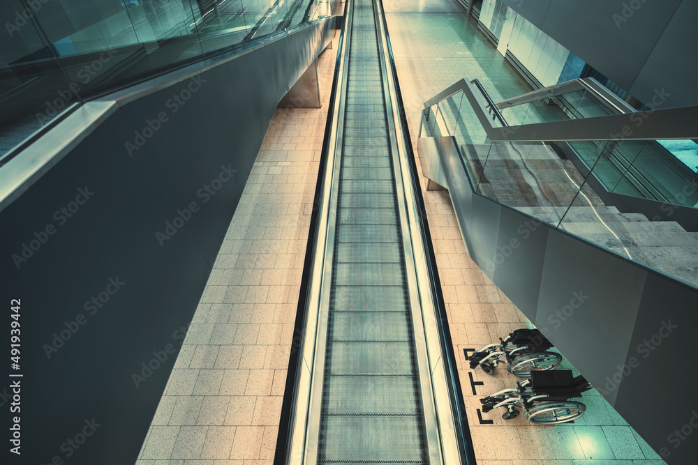 Obraz premium A view from above indoors of an airport or a railway station or a modern shopping: a long travelator, a moving staircase on the left and the stairway on the right; two wheelchairs under the stairs