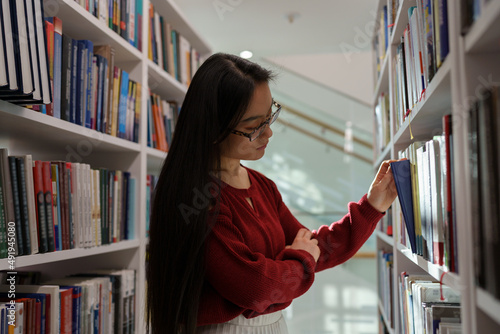 Thoughtful asian student girl searching materials for educational research in college library. Young chinese woman choosing book for reading in bookstore, selective focus. Literature and education