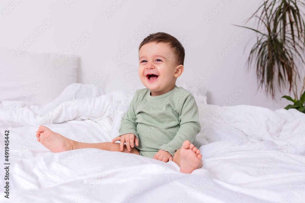 laughing baby in a green cotton bodysuit and bare feet is sitting on a bed spread out laughing baby in a green cotton bodysuit and bare feet is sitting on a bed spread out
