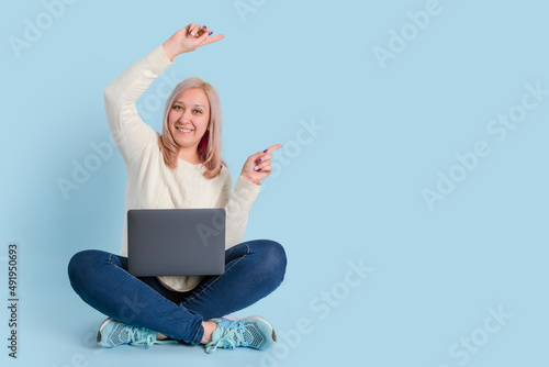 A woman sits with a laptop on her lap on a blue background and points to copy space with both hands © Larisa