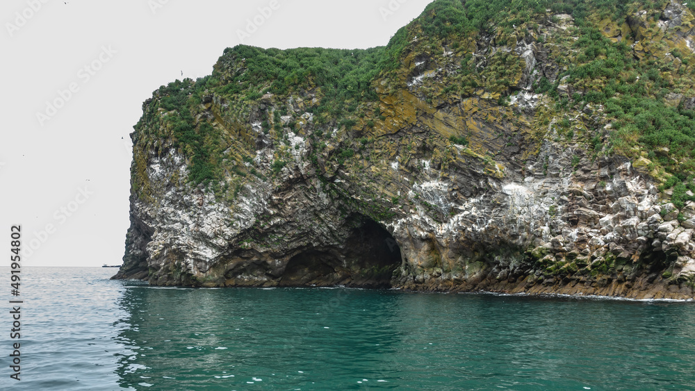 A rocky island in the Pacific Ocean. Green vegetation on steep slopes. A cave is visible above the water. Kamchatka. Avacha Bay