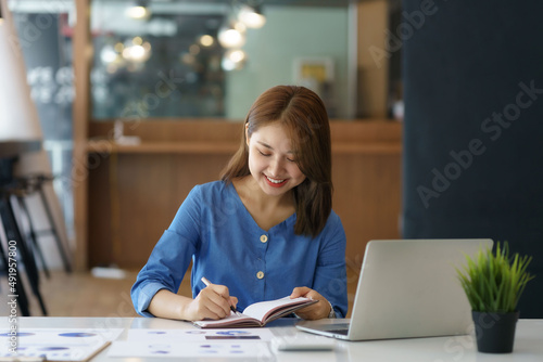 Charming Asian woman with a smile sitting and taking notes with computer laptop on her desk, enjoying work.