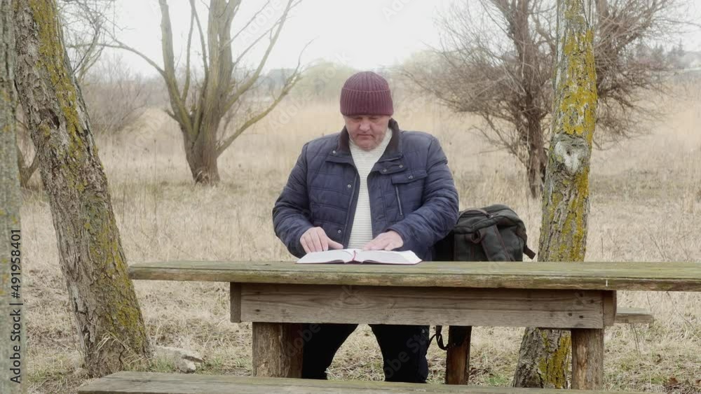 a man outdoors reads a book with interest sitting at a wooden table between trees a man outdoors reads a book with interest sitting at a wooden table between trees