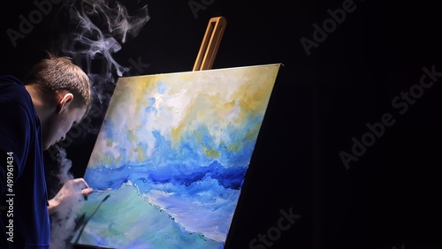 Artist copyist paint seascape with ship in ocean. Vaper smoke vape e-cigarette. Craftsman decorator draw as boat sail on blue sea with acrylic oil color. Draw finger, brush, knife palette. Indoor. photo