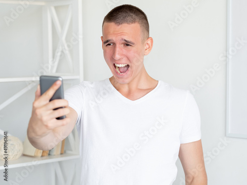 Screaming man. Virtual connection. Angry emotion. Mad stressed guy in white t-shirt looking smartphone holding hand in light room interior.