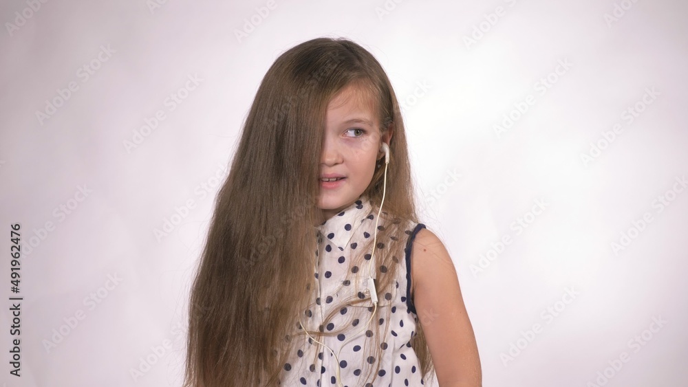 Child with earflaps dancing at studio background. The girl listens to music on the smartphone. The kid has loose long hair. Beautiful cute happy little girl listens to music on Headphones. Child with earflaps dancing at studio background. The girl listens to music on the smartphone. The kid has loose long hair. Beautiful cute happy little girl listens to music on Headphones.