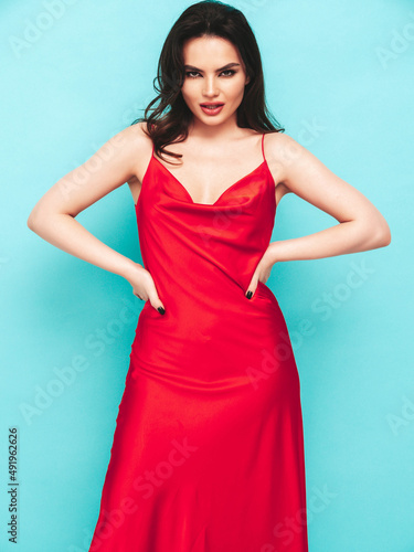 High fashion portrait of young beautiful brunette woman wearing nice trendy red evening dress. Sexy smiling fashion model posing in studio. Fashionable female isolated on blue