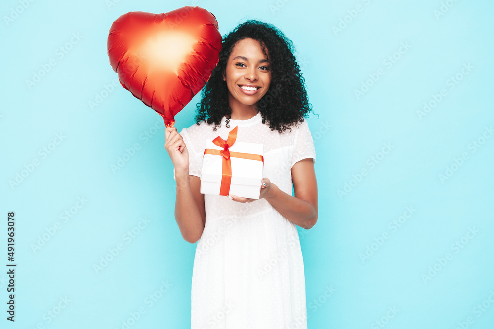 Beautiful black woman with afro curls hairstyle. Smiling model dressed in white summer dress. Sexy carefree female posing near blue wall in studio. Holding heart air balloon and gift box. Isolated