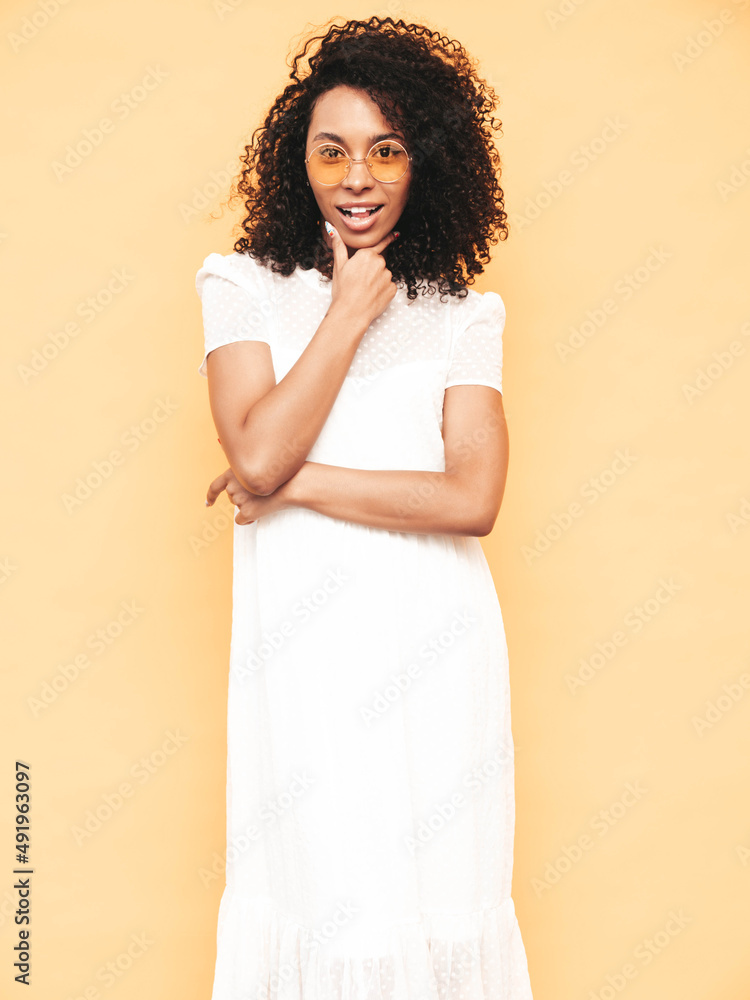 Portrait of beautiful black woman with afro curls hairstyle. Smiling model dressed in white summer dress. Sexy carefree female posing near yellow wall in studio. Tanned and cheerful. Isolated Portrait of beautiful black woman with afro curls hairstyle. Smiling model dressed in white summer dress. Sexy carefree female posing near yellow wall in studio. Tanned and cheerful. Isolated