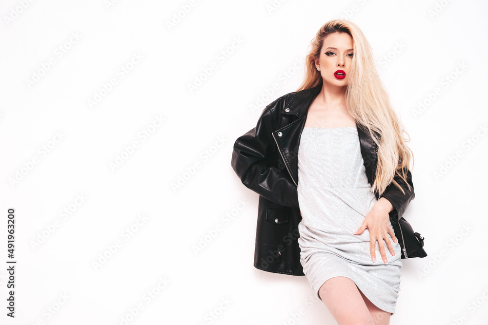Young beautiful confident female in trendy summer evening silver dress. Sexy carefree blond woman posing near white wall in studio. Model in black leather biker jacket and res lips Young beautiful confident female in trendy summer evening silver dress. Sexy carefree blond woman posing near white wall in studio. Model in black leather biker jacket and res lips