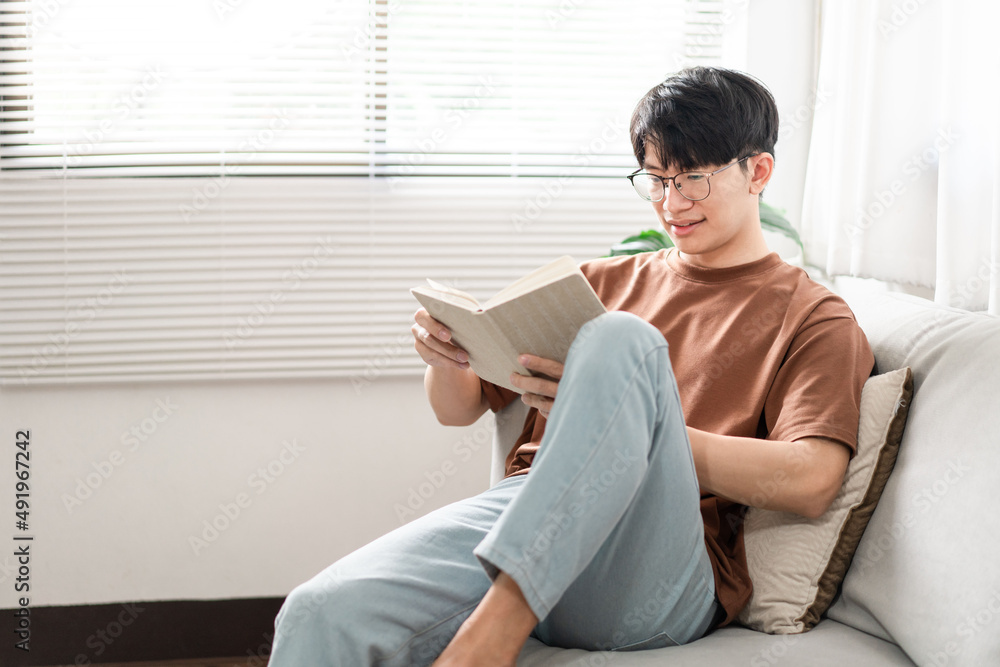Technology Concept The male with his glasses sitting on the grey sofa, leaning his back on it, and reading a book Technology Concept The male with his glasses sitting on the grey sofa, leaning his back on it, and reading a book