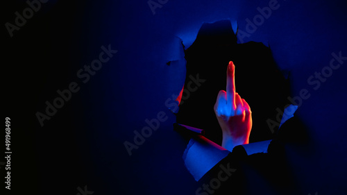 Fuck you. Provocative gesture. Red blue neon light female hand showing middle finger obscene sign inside breakthrough paper hole isolated on dark night copy space banner. photo