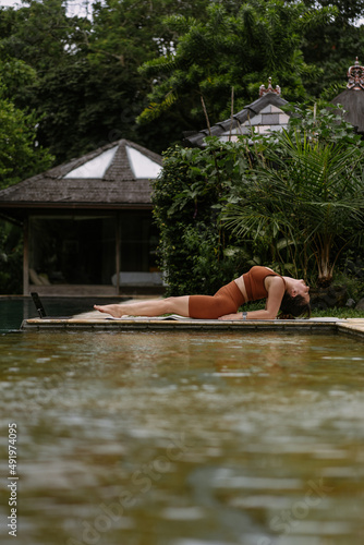Young woman with body positive appearance practicing yoga alone on deck by the pool in tropical island of Bali, Indonesia. Sport, fitness, healthy lifestyle concept.