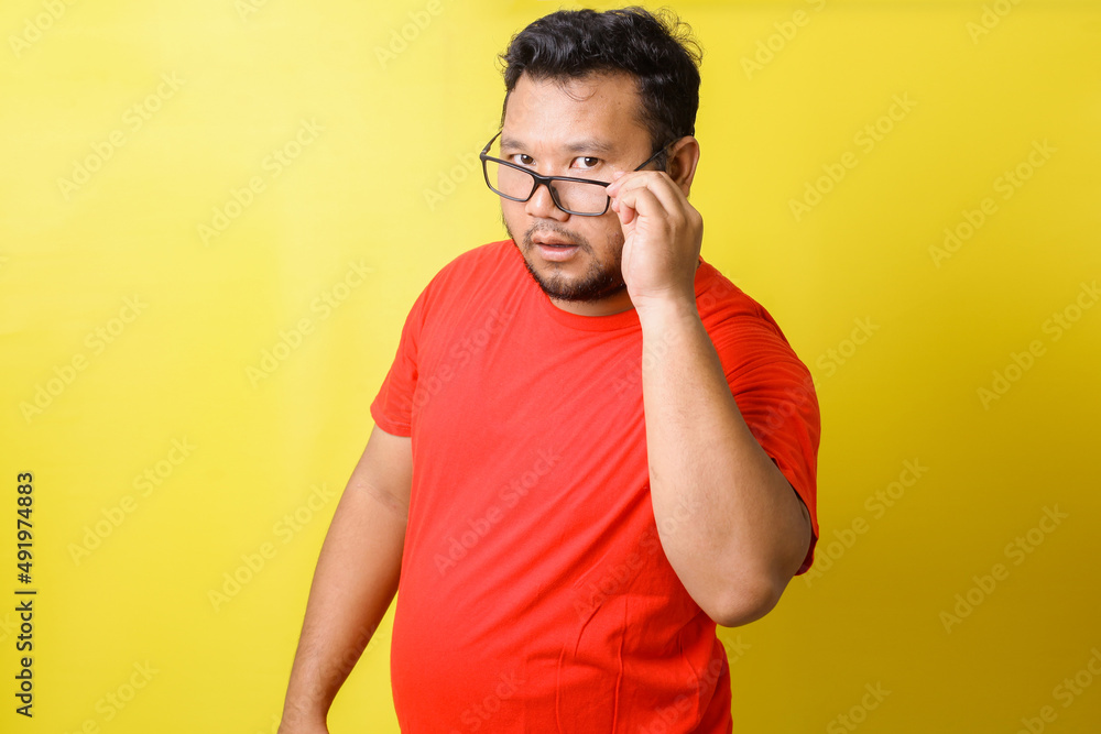 Asian fat man in red tshirt use eyeglasses is curious about something and looking seriously, isolated on yellow background Asian fat man in red tshirt use eyeglasses is curious about something and looking seriously, isolated on yellow background