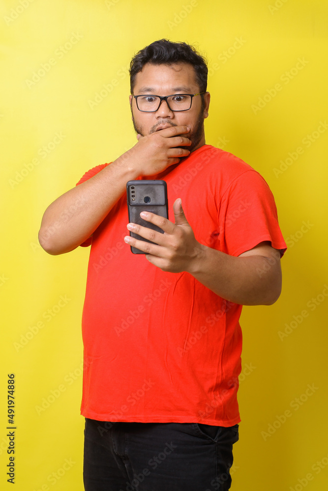 Fat asian guy with surprised expression covering mouth while looking to cell phone isolated on yellow background Fat asian guy with surprised expression covering mouth while looking to cell phone isolated on yellow background