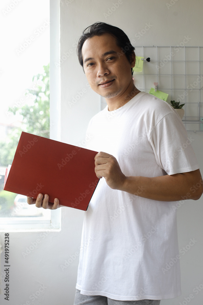 Asian man in white tshirt standing while holding a book with smiling expressions Asian man in white tshirt standing while holding a book with smiling expressions