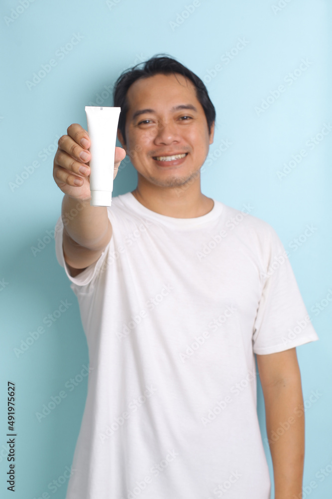 Smiling asian man showing plain tube isolated on pastel blue background. Tube mockup Smiling asian man showing plain tube isolated on pastel blue background. Tube mockup