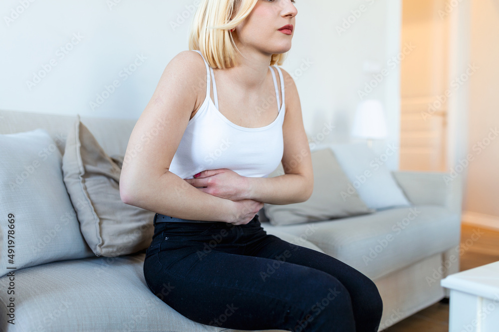 Woman in painful expression holding hands against belly suffering menstrual period pain, lying sad on home bed, having tummy cramp in female health concept