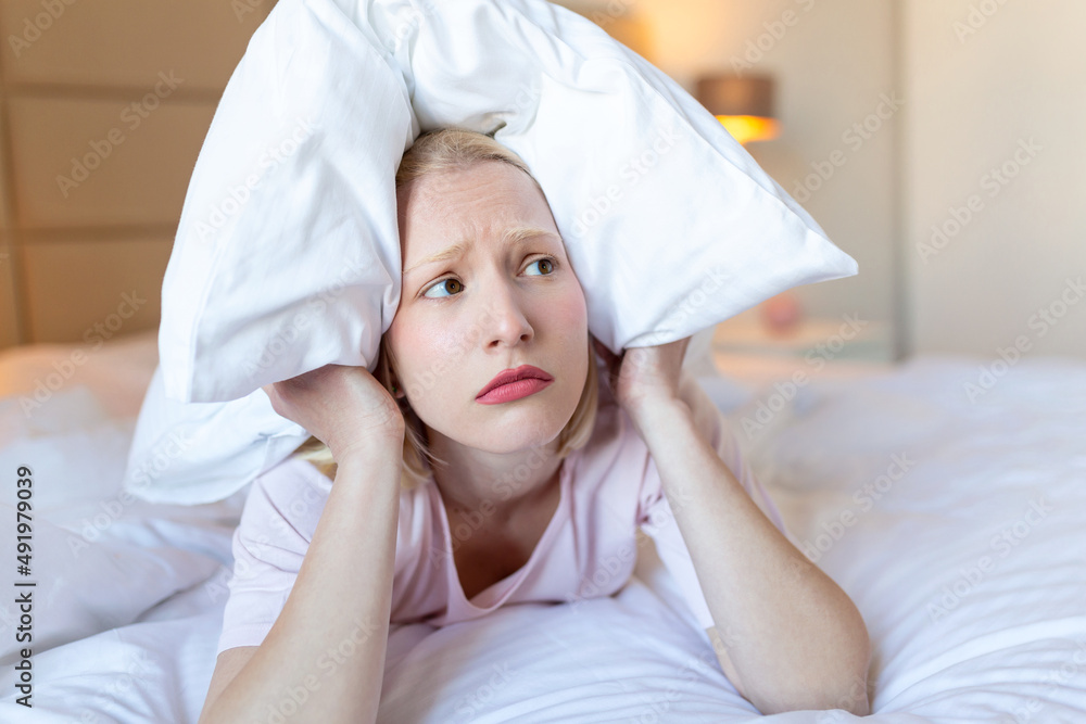 Angry woman suffering for neighbour noise lying on a bed in the night, covering ears with pillow because of noise. Angry woman suffering for neighbour noise lying on a bed in the night, covering ears with pillow because of noise.