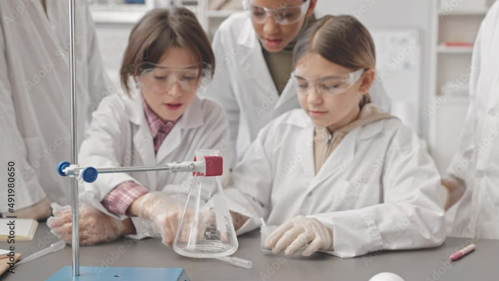 Medium slowmo of group of schoolkids in lab coats making chemical experiment by putting brown powder into glass flask with liquid and having lab smoke from flask as reaction Medium slowmo of group of schoolkids in lab coats making chemical experiment by putting brown powder into glass flask with liquid and having lab smoke from flask as reaction
