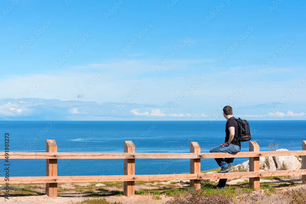Man with backpack sitting on the fence at the end of earth watching on bright blue lake. Hiking or travel concept. Exploring the great outdoors. Beauty of nature