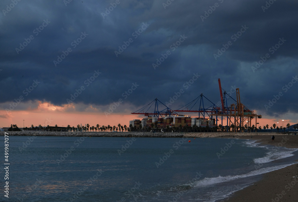 Colorful sunset over sea port and industrial cranes. Coastline of beach overlooking seaport of Spain. Transportation and logistic background - Sea containers are off loaded from vessels by huge cranes
