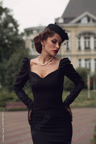 Retro portrait of beautiful European woman in black dress, with big red lips, retro hairdress and black hat with veil, Catholic Church in background © Tatyana