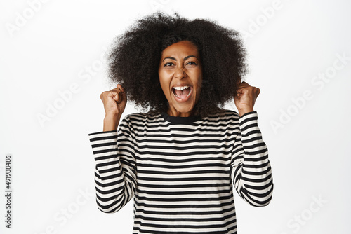 Enthusiastic african american woman celebrating, scream yes, looking amazed and surprised, winning, triumphing over success, standing against white background © Cookie Studio
