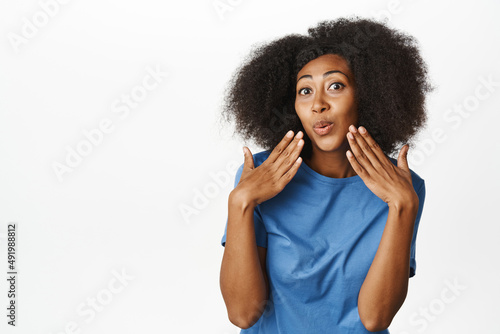 Portrait of coquettish female model with afro hair, chuckle and pucker lips silly, posing cute against white background in casual blue t-shirt