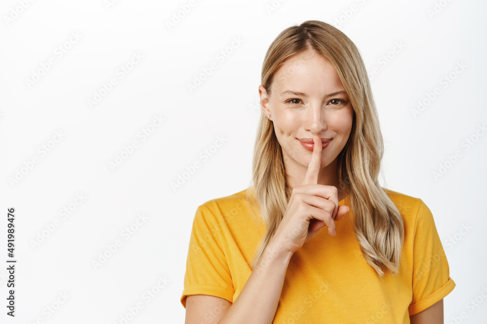 Smiling beautiful girl showing quiet, silence taboo sign, press finger to lips, hush sign, beauty secret concept, standing in yellow tshirt over white background Smiling beautiful girl showing quiet, silence taboo sign, press finger to lips, hush sign, beauty secret concept, standing in yellow tshirt over white background
