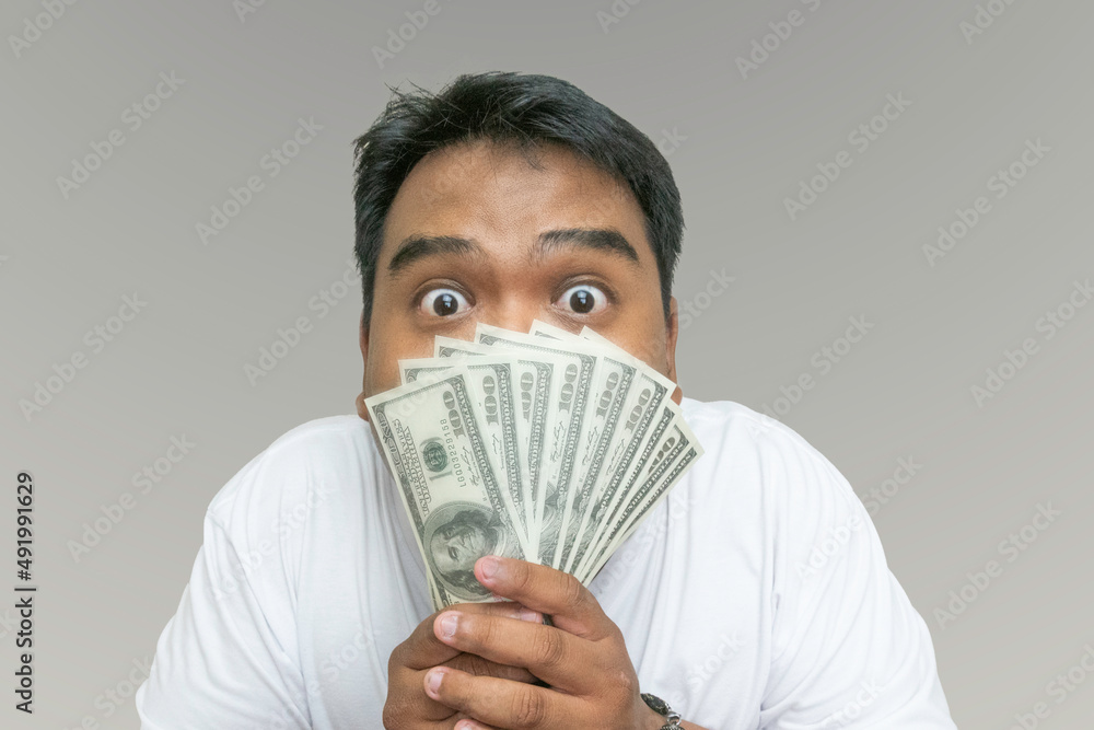 young man holding dollar money to cover his face with an excited face, big eyes over grey background. business, people, and finances concept. young man holding dollar money to cover his face with an excited face, big eyes over grey background. business, people, and finances concept.