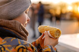 Kid eating boiled corn at the street in winter. Boy is wearing grey hat and overalls and holding corn. Vegetarian healthy food.