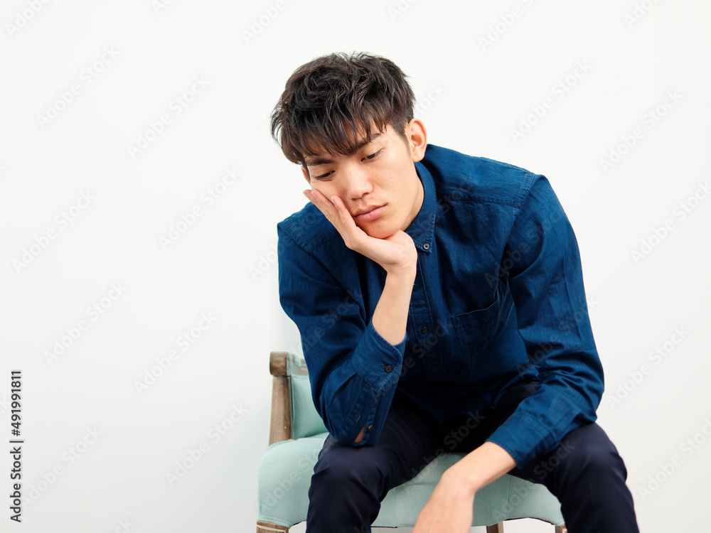 Portrait of handsome Chinese young man with curly black hair in blue shirt sitting in armchair and posing against white wall background. Hand on chin and looks sad, front view studio shot Portrait of handsome Chinese young man with curly black hair in blue shirt sitting in armchair and posing against white wall background. Hand on chin and looks sad, front view studio shot