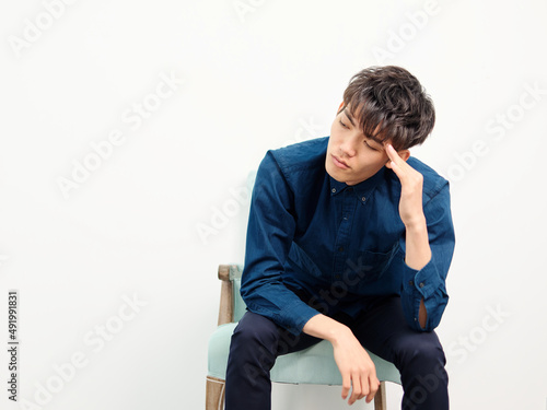 Portrait of handsome Chinese young man with curly black hair in blue shirt sitting in armchair and posing against white wall background. Fingers on forehead and looks tired, front view studio shot