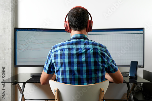 Back view of unrecognizable male employee in headphones reading graphs on computer screens while working in office